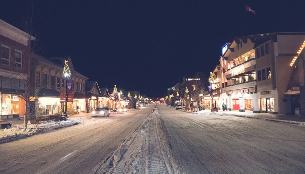 A photo of a snowy road in Banff, canada, with cars, store fronts and lights.