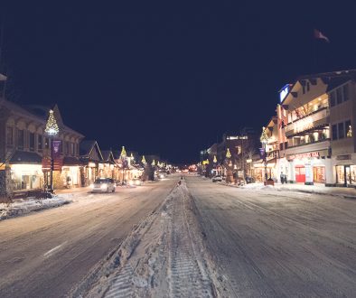 A photo of a snowy road in Banff, canada, with cars, store fronts and lights.